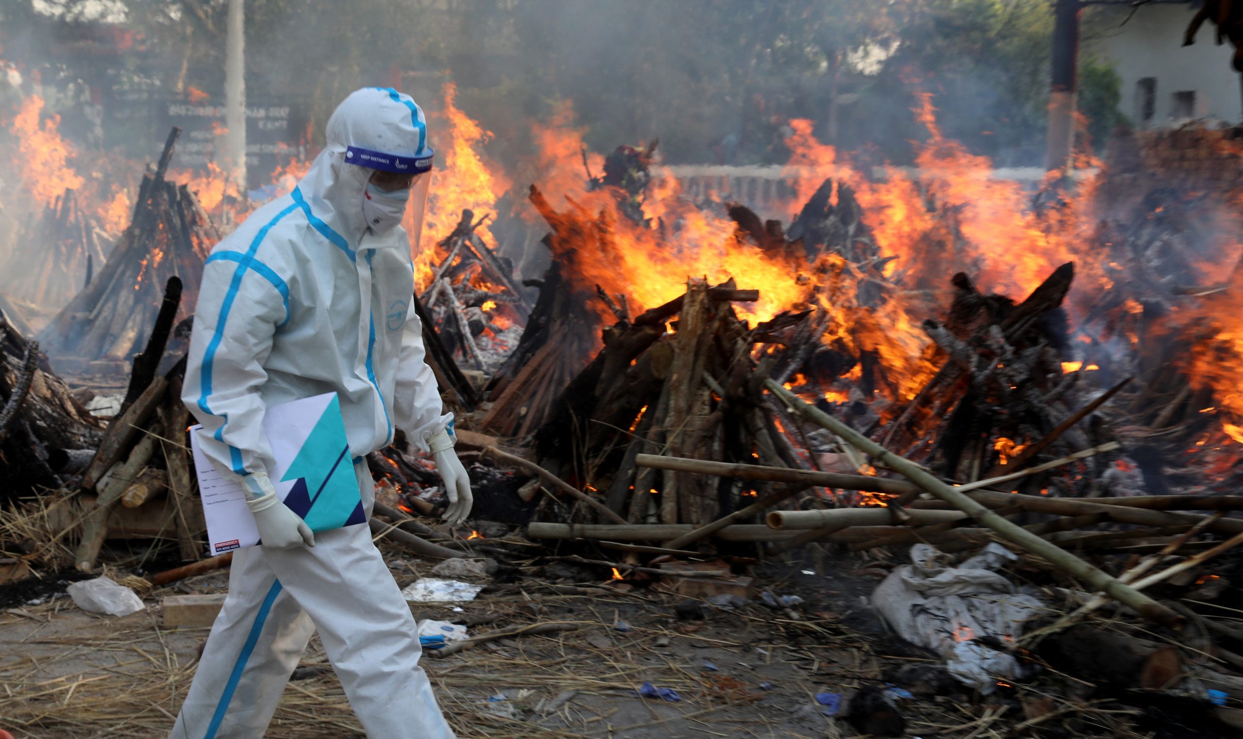 A man dressed in full PPE passing a mass cremation of Covid-19 victims in New Delhi, India, on April 30, 2021. Photo by Exposure Visuals/ Shutterstock.com.
