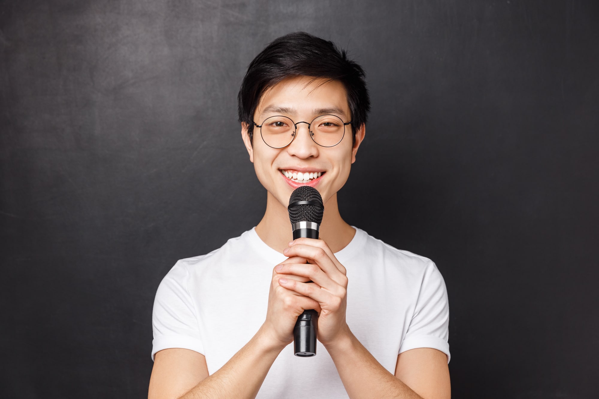 Leisure, people and music concept. Portrait of cute smiling asian man in white t-shirt, holding microphone both hands, singing song on karaoke party, perform in front of audience, black background